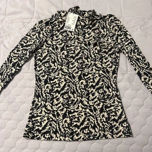 Mönstrad långärmad topp från H&M - Snygg långärmad topp från H&M med ett unikt svart och beige mönster. Perfekt för att ge din outfit en trendig touch. Toppen har en hög krage och är tillverkad i ett mjukt material.