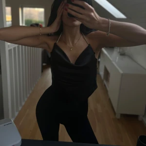 Svart satinlinne med strassdetaljer - Elegant svart satinlinne med tunna axelband dekorerade med strass. Toppen har en v-ringad design och knytning i ryggen för en justerbar passform. Perfekt för en stilren och chic look. Väl använd därför är strass detaljerna slitna