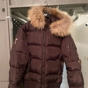 Brun dunjacka med päls från Bomboogie - Säljer en brun dunjacka från Bomboogie med stor päls på huvan. Jackan har en quiltad design och dragkedja framtill. Perfekt för kalla dagar med sin varma dunfyllning.              Nypris 2775 kr på Zalando