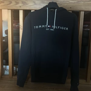 Svart hoodie från Tommy Hilfiger - Säljer en stilren svart hoodie från Tommy Hilfiger med logotyp och text 'EST. 1985' på bröstet. Pris kan diskuteras vid snabb affär