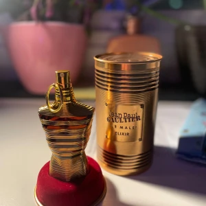 Jean Paul Gaultier Le Male Elixir Parfym - Elegant parfymflaska i form av en gyllene manlig torso från Jean Paul Gaultier. Flaskan står på en röd bas och har en matchande gyllene förpackning. Doften är en del av Le Male Elixir-serien och innehåller 75 ml. Perfekt för den som söker en lyxig och distinkt doftupplevelse.