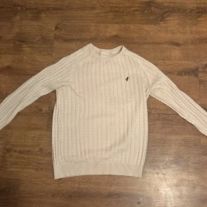 Vit stickad tröja från Pier One - Säljer en stilren vit stickad tröja från Pier One med ett diskret märke på bröstet. Tröjan har ett klassiskt flätmönster och ribbade muddar vid ärmslut och nederkant. Perfekt för kyligare dagar.