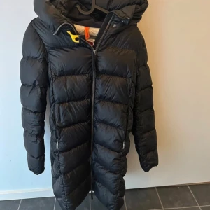 Svart dunjacka från Parajumpers - Säljer en svart dunjacka från Parajumpers med huva och dragkedja. Jackan har en logotyp på ärmen och är perfekt för kalla dagar. Den är lång och har en stilren design.