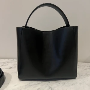 Svart handväska i skinn  - Elegant svart handväska i skinn med en stilren design. Väskan har ett robust handtag och en avtagbar axelrem för extra flexibilitet. Perfekt för en lite tråkigare look bättre. 