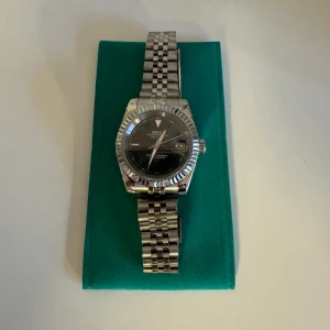 Seiko Wimbledon dial - Elegant silverfärgad klocka med en svart urtavla och datumvisning. Klockan har en klassisk länkarmband i rostfritt stål och en räfflad bezel. Perfekt för den stilmedvetna som vill ha en tidlös accessoar.