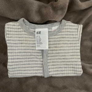 Babybody från H&M - Babybody från H&M i grå- och vitrandigt mönster. Bodyn har korta ärmar och knäppning.   Endast använt fåtal gånger. 