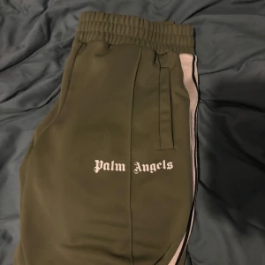 Gröna mjukisbyxor från Palm Angels - Snygga gröna mjukisbyxor från Palm Angels med elastisk midja och vita sidoremsor. Perfekta för en avslappnad stil. Byxorna har en broderad logga på sidan och är gjorda i ett bekvämt material.