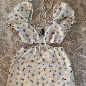 Kort kläning  - Vit kläning med blåa söta blommor, knytning fram och puffärmar, har ett innetyg i ”kjol” delen, används 1 gång och passar perfekt till sommaren eller avslutningar🩷