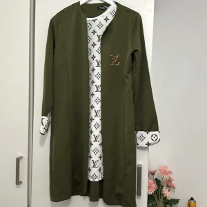 Olivgrön lång jacka med mönstrad insida - Säljer en olivgrön lång jacka med en cool mönstrad insida. Jackan har långa ärmar och en snygg detalj med ett glittrigt emblem på bröstet. Perfekt för att ge din outfit en unik touch!