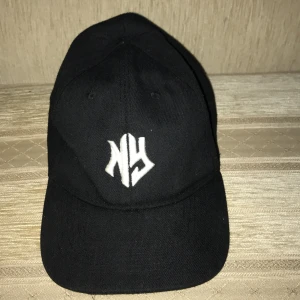 Svart keps med broderad logga - Säljer en stilren svart keps med en vit broderad 'NY' logga på framsidan. Perfekt för både vardag och sportiga tillfällen. Kepsen har en klassisk design med böjd skärm och justerbar rem baktill för bästa passform.