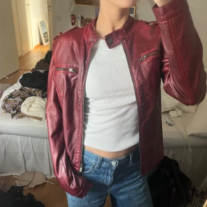 Röd skinnjacka - Säljer en snygg röd skinnjacka i nyskick. Den har en cool bikerstil med dragkedjor och en ståkrage. Perfekt för höst och vår, och funkar lika bra till fest som till vardags. Jackan har en normal passform och är långärmad. Den är gjord av skinn och har en härlig glansig finish.