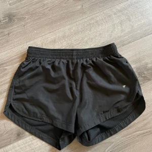 Svarta träningsshorts från Workout - Säljer ett par svarta träningsshorts från Workout i storlek XS. De är gjorda i ett lätt och bekvämt polyester-material, perfekt för träning. Shortsen har en elastisk midja för en bra passform och en liten logga på sidan. Inga hål och inga fläckar.