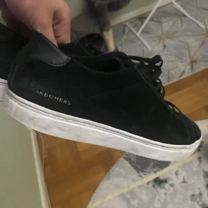 Svarta sneakers från Skechers - Säljer ett par svarta sneakers från Skechers. De är i mocka och har en klassisk design med snörning. Skorna har en vit sula som ger en snygg kontrast till det svarta. Perfekta för vardagsbruk och passar till det mesta i garderoben.