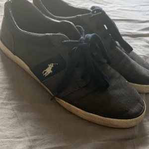 Svarta sneakers från Polo - Säljer ett par svarta sneakers från Polo. De har en klassisk design med snörning och en vit sula. Skorna är välanvända men fortfarande i bra skick. Perfekta för vardagsbruk!
