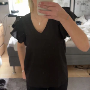 Svart blus från VILA - Säljer en elegant svart blus från VILA i storlek 34. Blusen har en v-ringad design och korta ärmar med volangdetaljer som ger en fin touch. Perfekt för både vardag och fest!