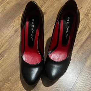 Svarta pumps från B&M Ricci - Säljer ett par Äkta läder! eleganta svarta pumps från B&M Ricci. De har en hög klack och en platåsula som ger extra höjd och stil. Skorna är gjorda i ett glansigt material med en röd insida som ger en lyxig känsla. Perfekta för festliga tillfällen!