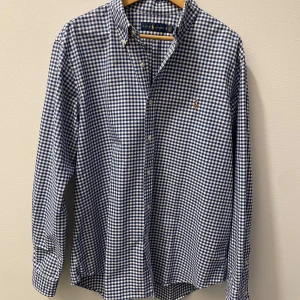 Blå och vit rutig skjorta från Ralph Lauren - Säljer en snygg blå- och vitrutig skjorta från Ralph Lauren. Skjortan har långa ärmar och en klassisk krage. Den är broderad med Ralph Laurens ikoniska logga på bröstet. Perfekt för både vardag och fest! Oanvänd i nyskick.