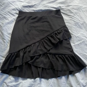 Svart kjol med volangdetaljer - Säljer en superfin svart kjol med volanger. Den har en omlott-look och är perfekt för både vardag och fest. Kjolen är ganska kort