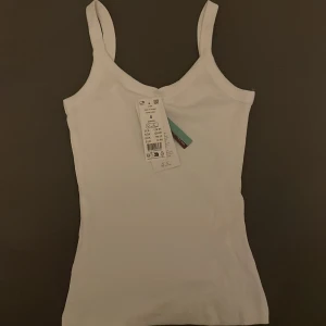 Vit ribbad linnetopp - Säljer en vit ribbad linnetopp i nyskick. Perfekt för sommardagar eller som ett basplagg i garderoben. Den är ärmlös med smala axelband och har en tight passform. Superbekväm och lätt att matcha med andra plagg!