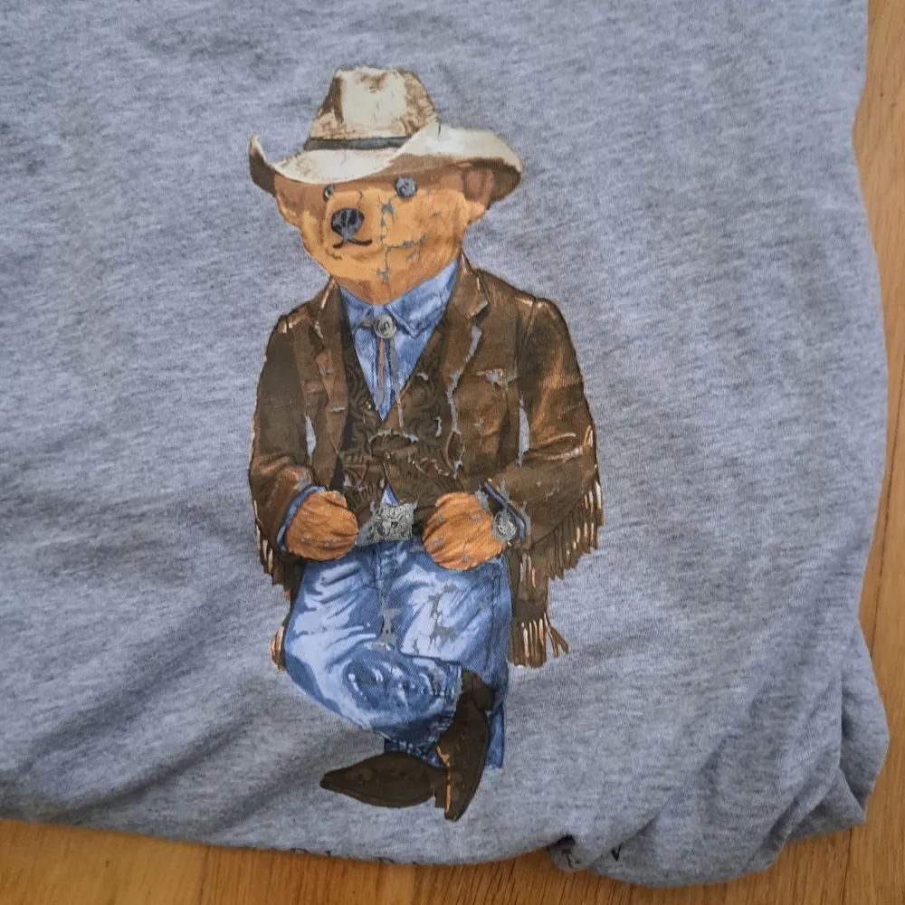 Säljer en grå t-shirt från Ralph Lauren med ett coolt tryck av en björn klädd i cowboy-outfit. T-shirten är perfekt för en avslappnad stil. Den är i okej skick med små defekter på etiketten och passar perfekt till vardags.. T-shirts.