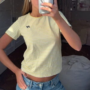 Gul- och vitrandig croppad t-shirt - Säljer en superfin gul- och vitrandig croppad t-shirt. Perfekt för sommardagar! Den har korta ärmar och en liten logga på bröstet. Passar perfekt till jeans eller shorts för en avslappnad look. 🐥