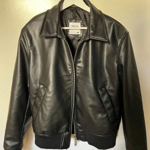 Svart skinnjacka från Zara - Säljer en snygg svart skinnjacka från Zara i storlek M. Jackan har en klassisk krage och dragkedja framtill. Perfekt för höst och vår, med ribbade muddar vid ärmslut och nederkant för en tight passform. Passar både till vardags och fest!