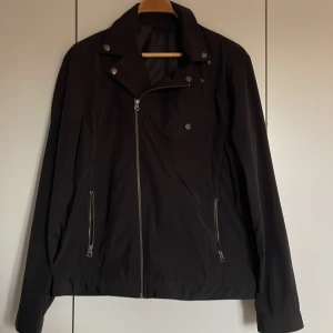 Svart jacka från Filippa K - Säljer en stilren svart jacka från Filippa K i storlek 54. Jackan har en asymmetrisk dragkedja och knappar vid kragen för en cool look. Den är långärmad och har två dragkedjefickor framtill. Perfekt för höst och vår!