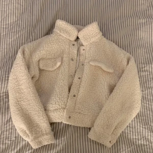 Vit teddyjacka - Mysig vit teddyjacka i nyskick. Den har en croppad passform med långa ärmar och knappar framtill. Perfekt för höst och vår när du vill hålla dig varm och stilren. Jackan har två fickor på bröstet för extra detaljer.