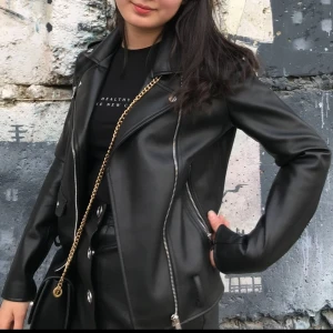 Svart skinnjacka från Zara - Säljer en snygg svart skinnjacka från Zara i storlek XS. Jackan har en cool bikerstil med dragkedjor och knappar som detaljer. Perfekt för höst och vår, och den ger en edgy touch till vilken outfit som helst. Jackan är i nyskick och redo för nya äventyr!