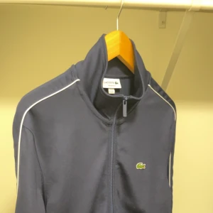 Mörkblå Lacoste Tröja - Tjena! Säljer en snygg mörkblå tröja från Lacoste i storlek M. Den har en dragkedja framtill och den klassiska Lacoste-loggan på bröstet. Jackan har långa ärmar med vita detaljer längs axlarna och ärmarna. Nu pris 1500-2000kr