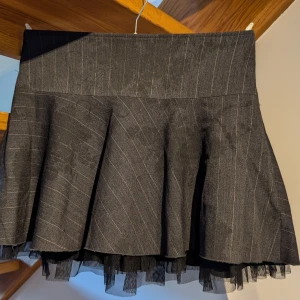 Kort vid kjol - Kort kjol från H&M. Mörk grå med tryckta stora svarta blommor och kritstrecksränder. Midjan 11 cm bred, total längd 36 cm. Underkjolen har svart tyll i nederkanten. Strl 36. Mkt fint skick.