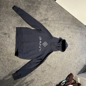 Mörkblå hoodie från GANT - Säljer en snygg mörkblå hoodie från GANT. Den har en stor logga på bröstet och coola detaljer med GANT-tryck på snörena. Perfekt för en avslappnad stil och passar till alla tillfällen. Den är i bra skick och superbekväm!
