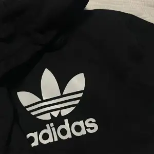 Säljer en svart hoodie från Adidas med den klassiska trefoil-loggan i vitt på framsidan. Den är perfekt för både vardag och träning. Hoodien har en bekväm passform och en stor ficka framtill. Perfekt för höst och vinter!