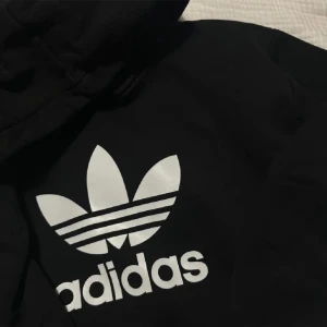 Svart hoodie från Adidas - Säljer en svart hoodie från Adidas med den klassiska trefoil-loggan i vitt på framsidan. Den är perfekt för både vardag och träning. Hoodien har en bekväm passform och en stor ficka framtill. Perfekt för höst och vinter!