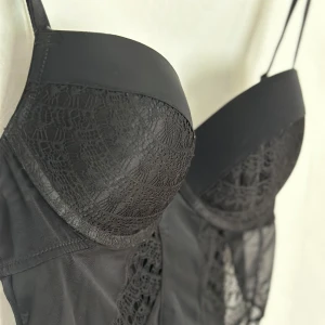 Svart korsett med spetsdetaljer - En elegant svart korsett med vackra spetsdetaljer i nyskick. Den har vadderade kupor (strl B/C)och justerbara axelband för perfekt passform. 