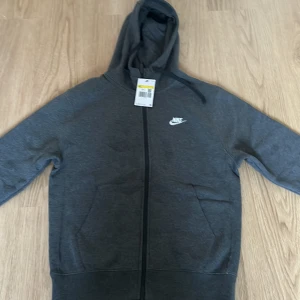 Grå hoodie från Nike - Säljer en snygg och bekväm grå hoodie från Nike. Den har en dragkedja framtill och en klassisk Nike-logga på bröstet. Perfekt för både vardag och träning. Den är i nyskick och redo för en ny ägare!