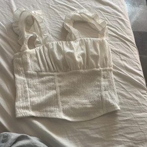 Vit topp från Gina Tricot - Säljer en superfin vit topp från Gina Tricot. Den är ärmlös med volangdetaljer på axelbanden och har en ribbad struktur. Perfekt för sommardagar eller en festlig kväll!
