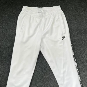 Vita mjukisbyxor från Nike - Säljer ett par vita mjukisbyxor från Nike i storlek S. Byxorna har en elastisk midja med snörning för bästa passform och sidofickor för praktisk förvaring. De har en stilren design med Nike-loggan på benet och en svart rand med text längs sidan. Perfekta för både träning och chill!
