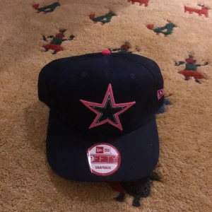 Svart keps med rosa stjärna - Snygg svart keps från New Era med en stor rosa stjärna broderad på framsidan. Kepsen har en snapback-stängning för justerbar passform och en rosa knapp på toppen. Perfekt för en cool och avslappnad stil.