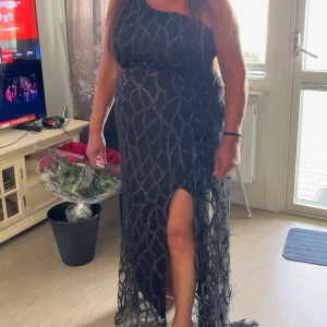 klänning  - Elegant svart oneshoulder klänning med ett unikt mönster och en slits framtill. Släpet på sidan har fjädrar & klänningen är i stretch material. 