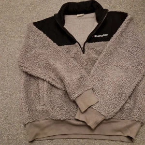 Grå fleece tröja från Champion - Mysig grå fleece tröja från Champion med svart panel upptill och dragkedja vid halsen. Perfekt för kyliga dagar med sin mjuka och varma känsla. Tröjan har långa ärmar och ribbade muddar vid ärmslut och nederkant.