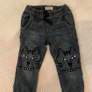 Blå jeansbyxor med kattmotiv från Lindex - Säljer ett par blå jeansbyxor från Lindex med coolt kattmotiv på knäna. Byxorna har en loose passform och elastisk midja med snörning för extra komfort. Perfekta för små äventyrare som vill ha stil och rörelsefrihet. Motivet på knäna gör det slitstarkt.