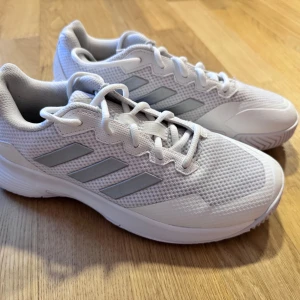 Vita sneakers från Adidas - Snygga vita sneakers från Adidas med klassiska tre ränder i silver på sidan. Skorna har en sportig design med snörning och en bekväm sula. Perfekta för en aktiv livsstil. Nypris: 1200 kr.