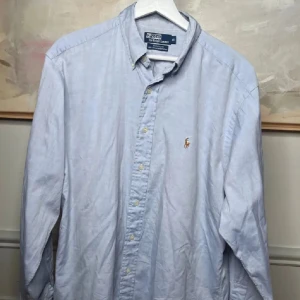 Ljusblå skjorta från Polo Ralph Lauren - Snygg ljusblå skjorta från Polo Ralph Lauren med klassisk krage och broderad logga på bröstet. Skjortan har långa ärmar och knappar framtill. Perfekt för en stilren look.