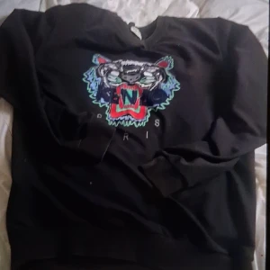 Svart tröja med tigertryck från Kenzo - Säljer en svart tröja från Kenzo med ett färgglatt tigertryck på framsidan. Tröjan har långa ärmar och en rund halsringning. Perfekt för en stilren och trendig look.