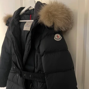 Svart dunjacka från Moncler - Snygg svart dunjacka från Moncler med pälsdetaljer på huvan. Jackan har ett bälte i midjan och dragkedja framtill. Perfekt för kalla dagar med sin långa design och varma material.