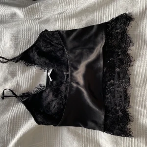 Svart satinlinne med spetsdetaljer - Snygg svart satinlinne med spetsdetaljer och smala axelband. Perfekt för en elegant look. Den har en v-ringad design och spetskant nedtill som ger en feminin touch.