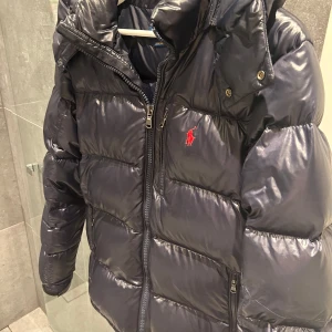 Mörkblå dunjacka från Ralph Lauren - Säljer en snygg mörkblå dunjacka från Ralph Lauren med en röd logotyp på bröstet. Jackan har en dragkedja framtill och en hög krage för extra värme. Perfekt för kyliga dagar! Riktigt bra skick 