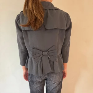 Grå kavaj med rosett - Snygg grå kavaj med trekvartsärm och dubbelknäppning. Den har en stor dekorativ rosett på ryggen och volangdetaljer nedtill. Perfekt för en stilren look.