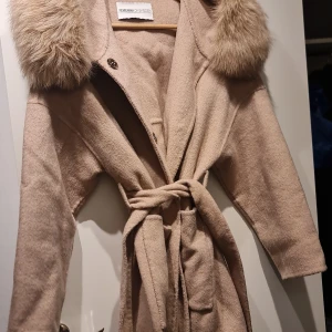 Beige kappa med fuskpäls från Femenina of sweden  - Elegant beige kappa med pälsdetaljer på kragen från Femenina of sweden . Kappan har ett knytbälte i midjan och långa ärmar. Perfekt för kyligare dagar med sin stilrena design och mjuka material.